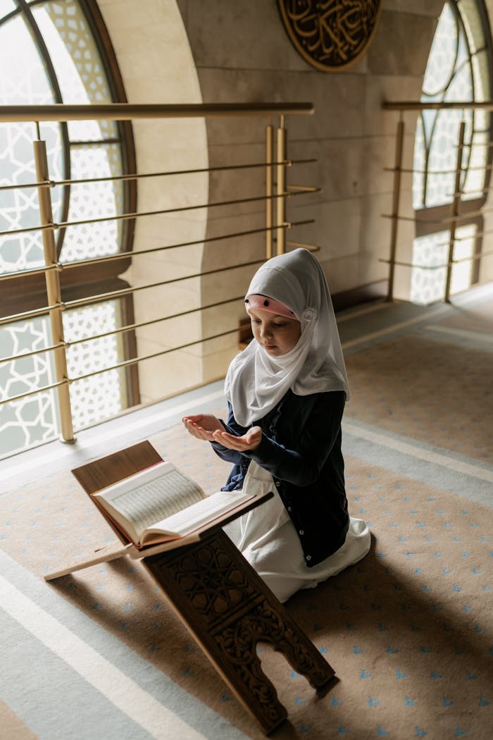 why-choose-us A young Muslim girl in a hijab prays indoors beside an open Quran on a stand.